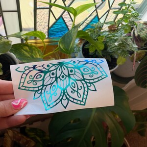 May include: A teal and blue iridescent vinyl decal of a half mandala design. The decal is on a white backing paper.