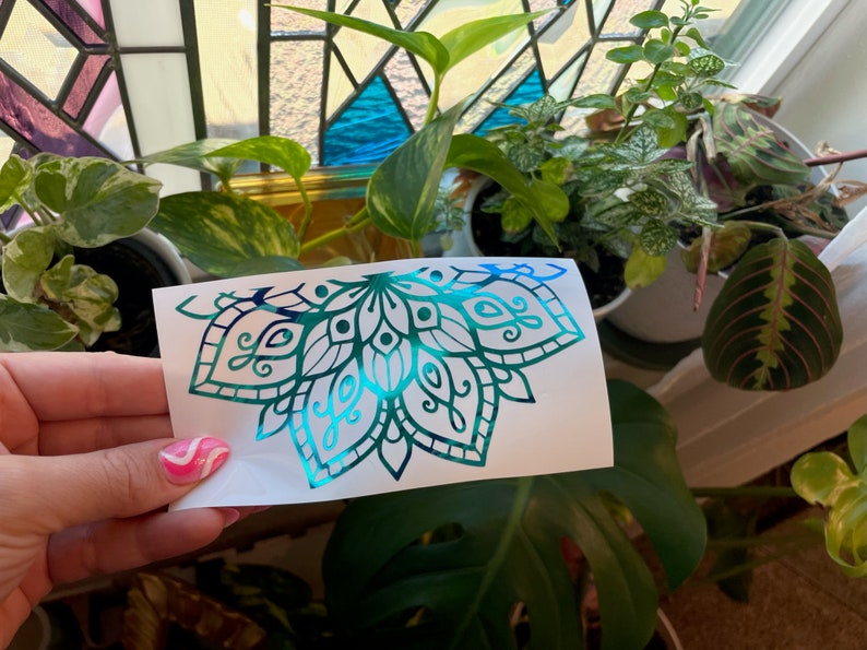 May include: A teal and blue iridescent vinyl decal of a half mandala design. The decal is on a white backing paper.