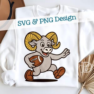 Puede incluir: Sudadera blanca con un carnero de dibujos animados sosteniendo un balón de fútbol americano. El carnero es gris con cuernos color beige y zapatos marrones. El texto "SVG & PNG Design" está impreso en la parte superior.
