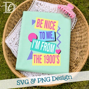 May include: A pale green t-shirt featuring the text "BE NICE TO ME, I'M FROM THE 1900'S" in a vibrant, retro style. The shirt is folded in a wicker basket with a white lace doily and a pink hair clip.