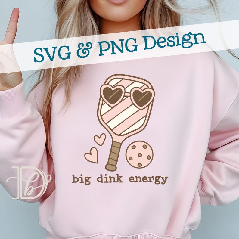 May include: Pink sweatshirt with a pickleball paddle design. The paddle has heart-shaped sunglasses and is paired with a pickleball and two hearts. The text "big dink energy" is below the design. The words "SVG & PNG Design" are at the top.