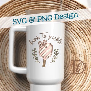 May include: White travel mug with a handle, featuring the text "born to pickle" in a script font. The design includes a pickleball paddle with pink and white stripes, a bow, and laurel branches. The words "SVG & PNG Design" are at the top.
