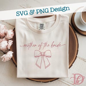 May include: Cream-colored t-shirt with the text "mother of the bride" in a cursive font, along with a bow graphic. The shirt also has the text "SVG & PNG Design" at the top. The shirt is displayed on a wooden surface.
