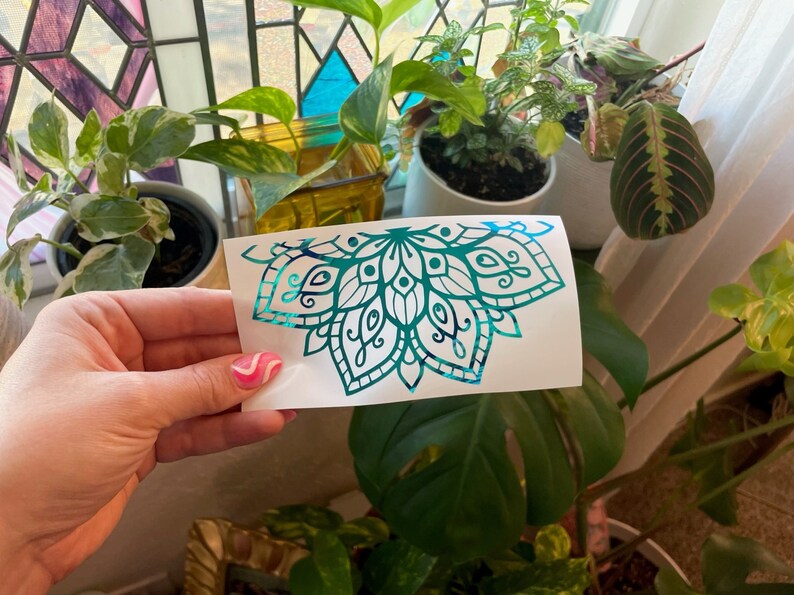 May include: A teal and blue iridescent vinyl decal of a mandala design. The decal is on a white background and is being held by a hand.