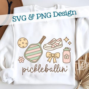 May include: White sweatshirt with a "pickleballin'" design. The design includes a pickleball paddle, ball, shoe, water bottle, bow, and stars in pastel colors. Text at the top reads "SVG & PNG Design".