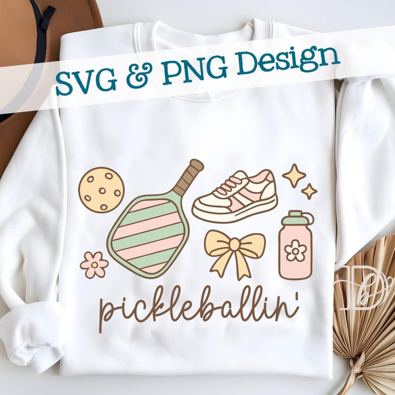 May include: White sweatshirt with a "pickleballin'" design. The design includes a pickleball paddle, ball, shoe, water bottle, bow, and stars in pastel colors. Text at the top reads "SVG & PNG Design".