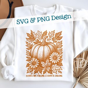 Pode incluir: Camisola branca com um design de outono. O design apresenta uma abóbora, folhas e flores em tons de laranja e castanho. O texto "SVG & PNG Design" está na parte superior e "LEAVES ARE FALLING & COZY IS CALLING" na parte inferior.