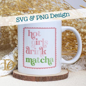 May include: White ceramic mug with the text "hot girls drink matcha" in pink and green, framed by a pink scalloped border. The mug sits on a small wooden coaster, with a blurred background of dried flowers.