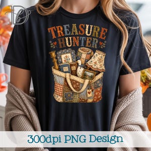 Puede incluir: Una camiseta negra con un estampado gráfico que incluye el texto "TREASURE HUNTER" sobre un bolso de retazos lleno de varios objetos, como un candelabro de latón, libros, una pequeña figura de pato, una lámpara y cuadros.
