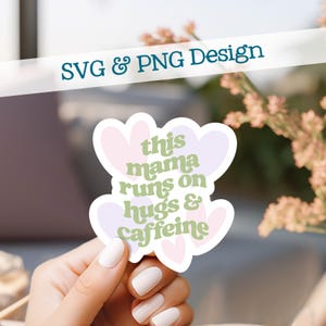 Könnte beinhalten: Ein weißer Aufkleber mit herzförmigem Design und dem Text "this mama runs on hugs & caffeine" in Grün und Lila. Der Aufkleber wird von einer Hand mit weißem Nagellack gehalten, mit den Worten "SVG & PNG Design" im Hintergrund.