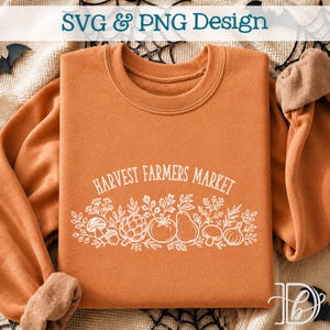 May include: A burnt orange crewneck sweatshirt featuring a white line art graphic of various vegetables and fruits, including a mushroom, artichoke, tomato, pear, and garlic, with the text Harvest Farmers Market arched above the produce.