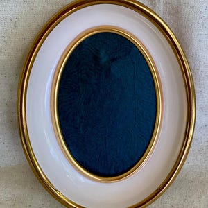 May include: An oval picture frame featuring a shiny gold outer rim, a smooth white inner border, and a thin gold inner edge surrounding a dark teal fabric center with a subtle, wavy, embossed texture throughout the fabric surface.