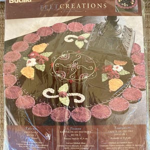 May include: A Bucilla Felt Creations doily kit. The kit includes a round felt doily with a floral design, featuring red, white, and yellow accents. The doily is surrounded by maroon felt circles. The kit is designed by Rebekah Meier.