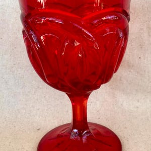 May include: A vibrant red glass goblet with a detailed, textured design. The goblet features a rounded bowl, a slender stem, and a circular base. The glass has a glossy finish, reflecting light.