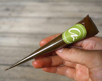 May include: A brown henna cone with a green label that reads "aminamalikhenna Eucalyptus Blend". The cone is held in a hand against a light wood grain background. The henna is a dark brown color.