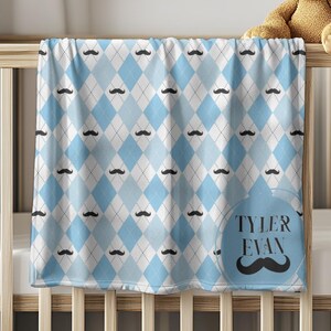 Op de afbeelding: Een babydeken met een blauw en wit ruitpatroon en zwarte snoraccenten. De deken ligt over een houten wieg. Een teddybeer zit op de rand van de wieg. De tekst "TYLER EVAN" staat in een blauwe cirkel.