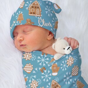 Könnte beinhalten: Eine blaue und weiße Wickeldecke mit einem Lebkuchenhaus, Schneeflocken und dem Namen "Oliver" darauf gedruckt. Ein Baby schläft in der Wickeldecke und trägt eine passende blaue und weiße Mütze mit dem Namen "Oliver" darauf.