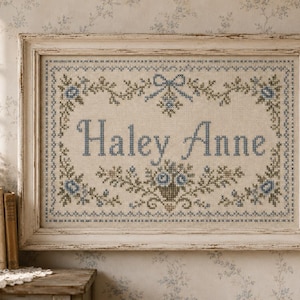 May include: A rectangular cross-stitch embroidery piece featuring the name Haley Anne in blue thread, centered within a decorative border of blue flowers and green vines, housed in a distressed off-white wooden frame with ornate molding.