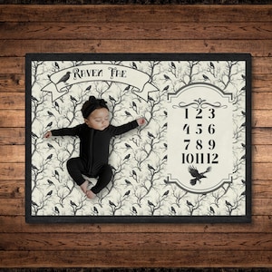 Könnte beinhalten: Eine weiße Babydecke mit einem schwarz-weißen Raben- und Baumdesign. Die Decke hat ein Banner mit der Aufschrift "Raven Tree" und eine nummerierte Tabelle von 1 bis 12. Ein Baby liegt auf der Decke.