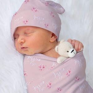 Op de afbeelding: Een roze swaddle-deken met een bloemenpatroon en de naam "Jessica Lynn" die overal wordt herhaald. Een baby slaapt in de swaddle, met een bijpassende roze muts met hetzelfde bloemenpatroon en dezelfde naam.