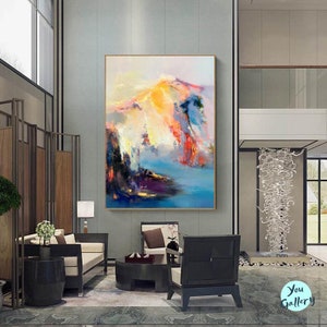 May include: Abstract painting with a colorful landscape scene. The painting features a mix of blues, pinks, yellows, and oranges. The painting is framed in a gold frame and is hanging on a wall in a modern living room.
