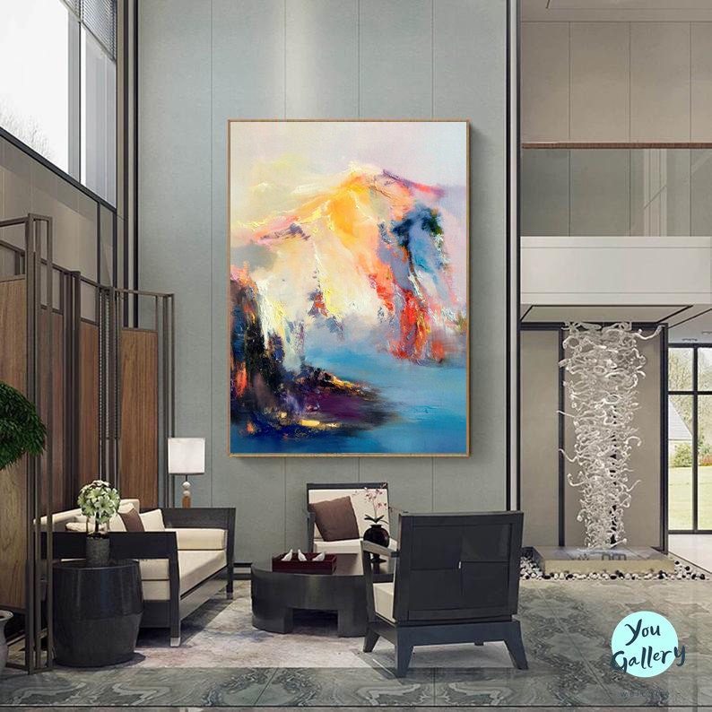 May include: Abstract painting with a colorful landscape scene. The painting features a mix of blues, pinks, yellows, and oranges. The painting is framed in a gold frame and is hanging on a wall in a modern living room.