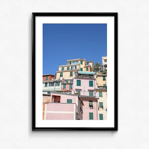 May include: A photograph of a row of colourful houses stacked on top of each other against a bright blue sky. The houses are painted in shades of pink, yellow, and orange, with green shutters and balconies.