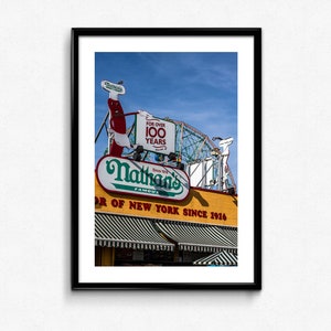 以下が含まれることがあります： 「Nathan's Famous」と「Flavor of New York Since 1916」と書かれた黄色と緑の看板。看板の上には、赤と白のホットドッグのキャラクターが描かれている。ホットドッグのキャラクターの後ろには、「For Over 100 Years」と書かれた白い看板が見える。