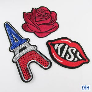 May include: Three embroidered iron-on patches. One is a red rose, one is a red and blue Eiffel Tower, and one is a red pair of lips with the word "KISS" in white.