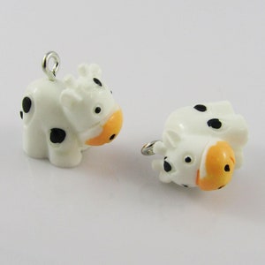 May include: Two white plastic cow charms with black spots and orange noses. The charms have silver jump rings for attaching to jewellery or other crafts.