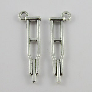 May include: Two silver-coloured crutch charms. Each charm features a loop at the top for attachment. The crutches have a detailed design, with vertical supports and a lower section, suitable for jewellery making.