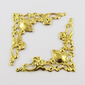 May include: Two gold metal corner embellishments with a decorative floral design. The embellishments are ornate and have a detailed, intricate design.
