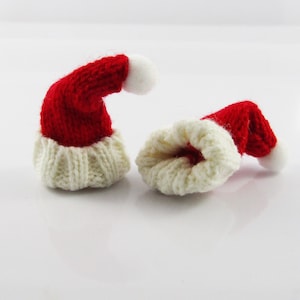 Peut inclure: Deux petits bonnets de Noël en tricot, l'un rouge avec une bordure blanche et un pompon blanc, l'autre retourné pour montrer la doublure en tricot blanche.