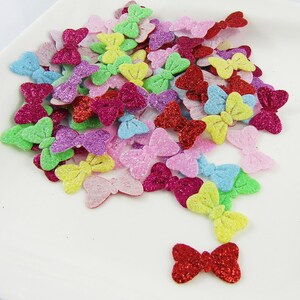 May include: A large pile of colourful glitter bows. The bows are made of felt and have a variety of colours, including red, pink, purple, blue, green, yellow, and orange.