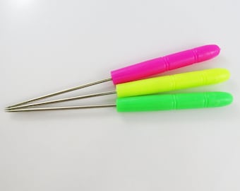 May include: Three colorful awls with stainless steel needles. The handles are bright pink, neon yellow, and green. The awls are arranged in a fan shape, with the needle tips pointing towards the left.