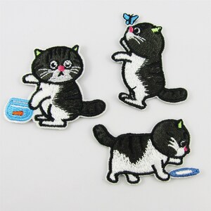 May include: Three black and white embroidered iron-on patches of cats. One cat is holding a fishbowl, another is chasing a butterfly, and the third is holding a blue ribbon.