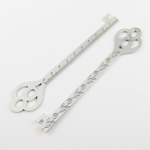 May include: A silver key with a decorative floral design and a ruler on the back. The ruler measures up to 25.4 cm.