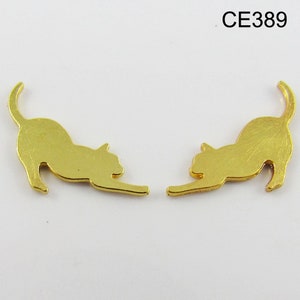 Puede incluir: Un par de pendientes de color dorado con forma de silueta de gato. Los gatos se miran, con sus colas enroscadas alrededor de sus cuerpos. Los pendientes tienen un dise&ntilde;o simple y elegante.