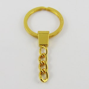 May include: A gold-coloured keychain featuring a large circular ring, a rectangular connector, and a short chain of three oval links. The keychain is made of shiny metal against a white background.