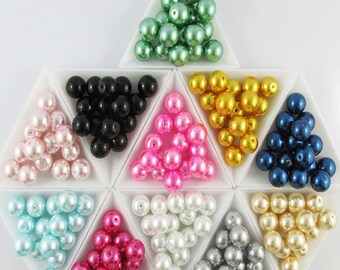 May include: Assortment of pearl beads in various colors, including pink, white, black, blue, green, gold, and silver. The beads are arranged in triangular compartments on a white surface.