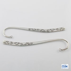 May include: Two silver metal bookmarks with a floral design. Each bookmark has a curved hook at one end and a small hole. The floral pattern is etched along the length of the bookmark.