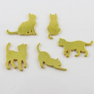 Puede incluir: Cinco amuletos de metal dorado en forma de silueta de gato. Los amuletos est&aacute;n en varias poses, incluyendo sentados, de pie y acostados.