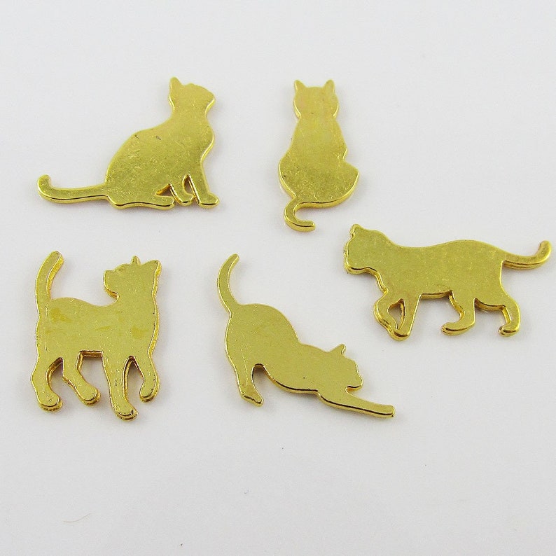 Puede incluir: Cinco amuletos de metal dorado en forma de silueta de gato. Los amuletos est&aacute;n en varias poses, incluyendo sentados, de pie y acostados.