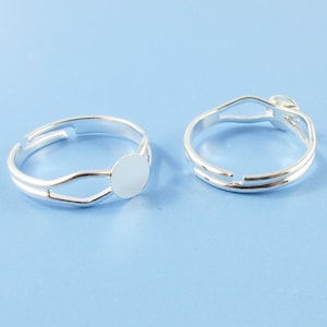 May include: Two silver adjustable rings with a round bezel for a cabochon. The ring on the left has a bezel that is open on one side. The ring on the right has a bezel that is closed on all sides.