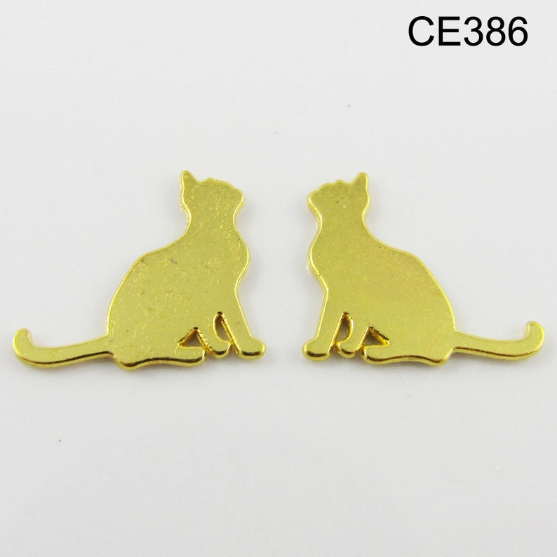 Puede incluir: Dos abalorios de metal de color dorado en forma de silueta de gato. Los gatos est&aacute;n sentados con las colas enroscadas alrededor de sus cuerpos.