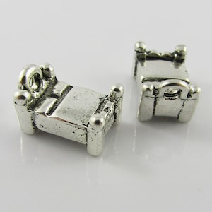 May include: Two silver charm pendants shaped like beds with a headboard and footboard. The charms have a loop at the top for attaching to a bracelet or necklace.