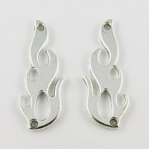Op de afbeelding: Twee zilverkleurige, vlamvormige bedels. Elke bedel heeft een gladde, metallic afwerking met uitgesneden details en een klein gaatje aan de boven- en onderkant om aan sieraden of andere knutselwerken te bevestigen. De bedels zijn ontworpen om op gestileerde vlammen te lijken.