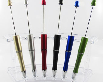 Puede incluir: Seis bolígrafos retráctiles en un soporte de plástico transparente. Los bolígrafos son dorados, plateados, rojos, negros, azules y verdes.