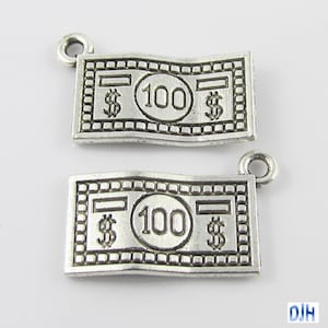 May include: Two silver-toned metal charms shaped like dollar bills. Each charm features the text "$100$" and a loop for attaching to a necklace or bracelet.