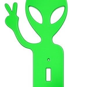 May include: A bright green alien-shaped light switch cover. The alien is depicted with large oval eyes and is giving a peace sign with one hand. The cover has a rectangular opening for the switch and two screw holes.